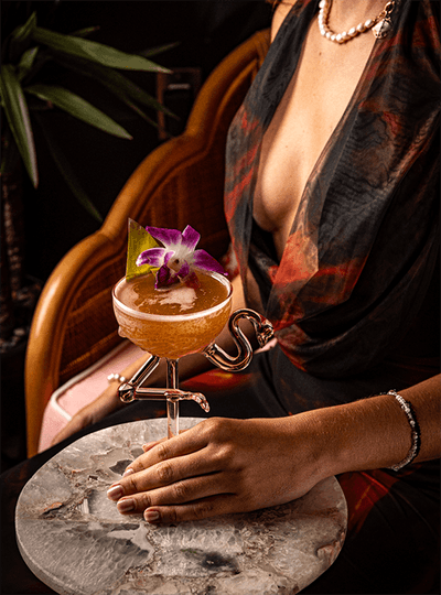 Lady holding a cocktail garnished with an orchid, resting on a sculptural stand and marble table at Esme Miami Beach