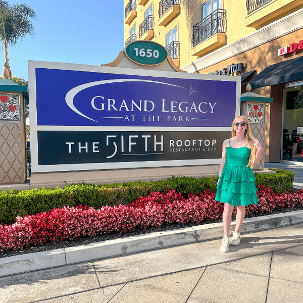 Guest in a green dress standing beside the Grand Legacy At The Park entrance sign on Harbor Boulevard in Anaheim under a clear blue sky.
