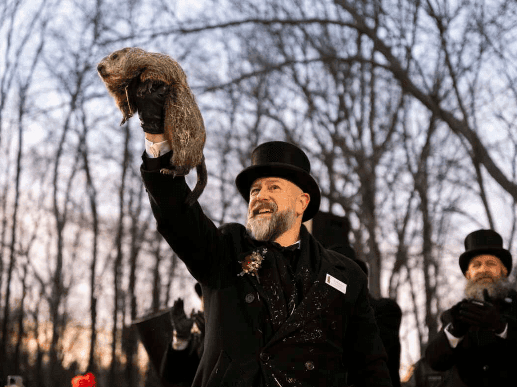 Man in top hat and black coat holds a groundhog aloft in winter woods for Groundhog Gets Cozy offer.