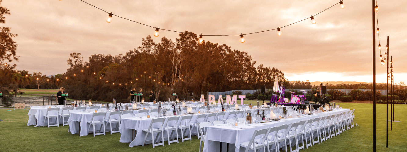 Outdoor event setup with long tables, white chairs, and string lights over a grassy area at dusk.