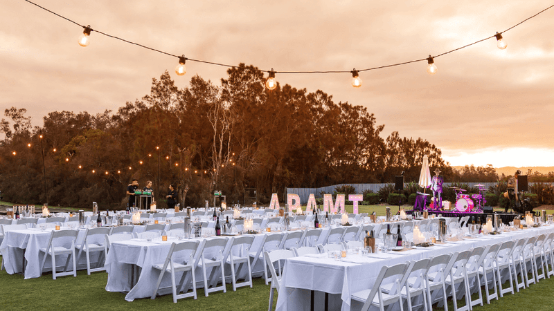 Outdoor event setup with long tables, white chairs, and string lights over a grassy area at dusk.