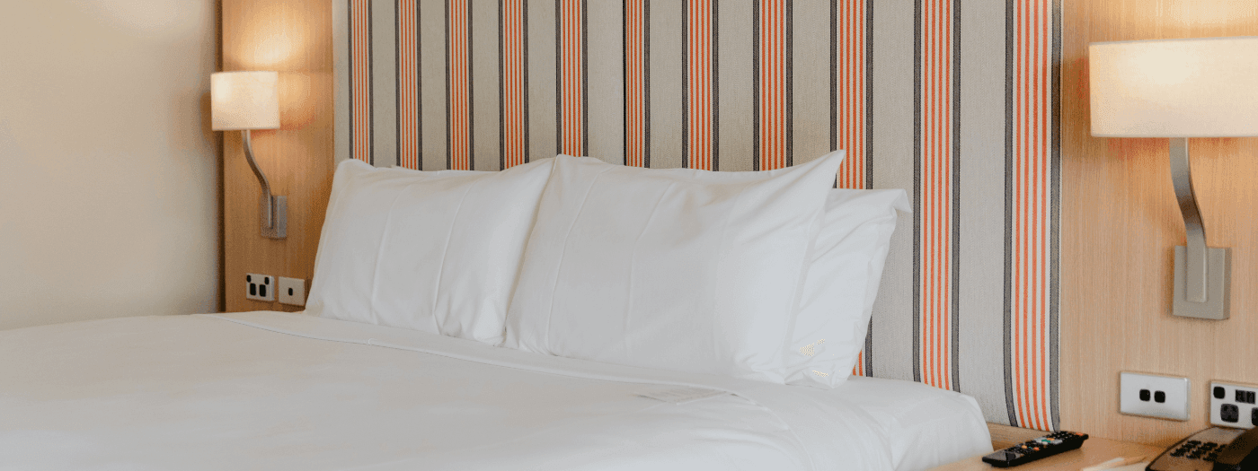Hotel room with a bed, two pillows, wooden nightstand, telephone, remote, and two lamps.