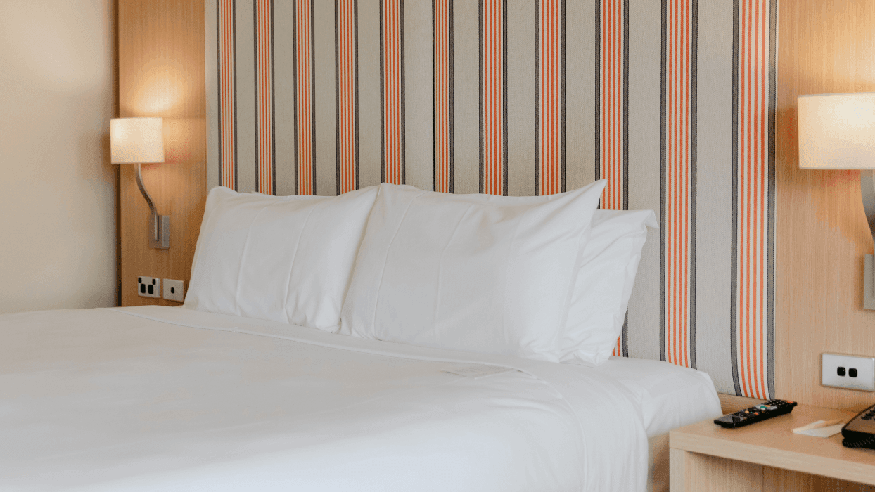 Hotel room with a bed, two pillows, wooden nightstand, telephone, remote, and two lamps.