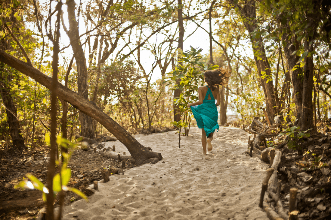 Woman in a teal dress runs through a sun-drenched, sandy forest path near Cala Luna Boutique Hotel