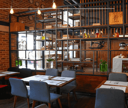 Industrial dining room with wooden tables with chairs under exposed bulbs before an open metal shelf at Unika Hotels