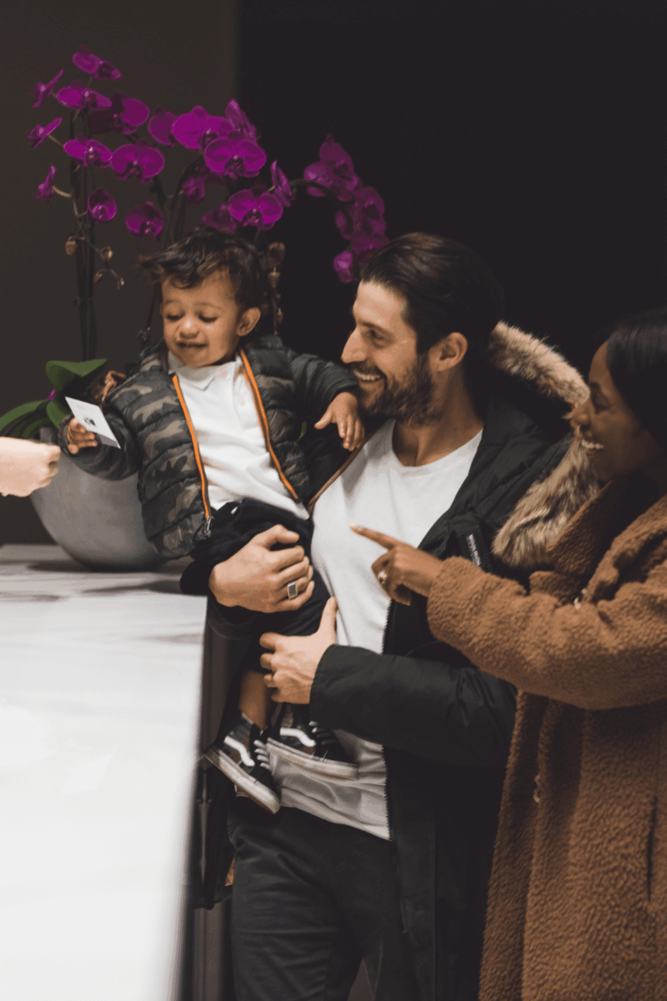 Father holding a toddler by a check-in desk with purple orchids, near a smiling woman at Hotel X Toronto