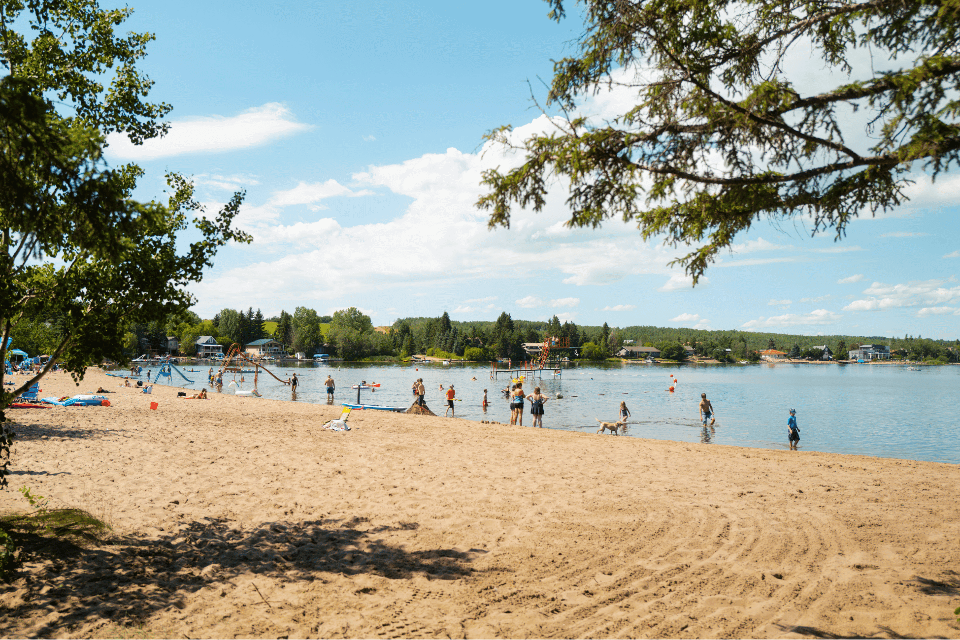Photograph of a sandy lakeside beach with people swimming, walking, and relaxing under a partly cloudy sky. Trees frame the scene on both sides, with houses and greenery visible across the calm water.