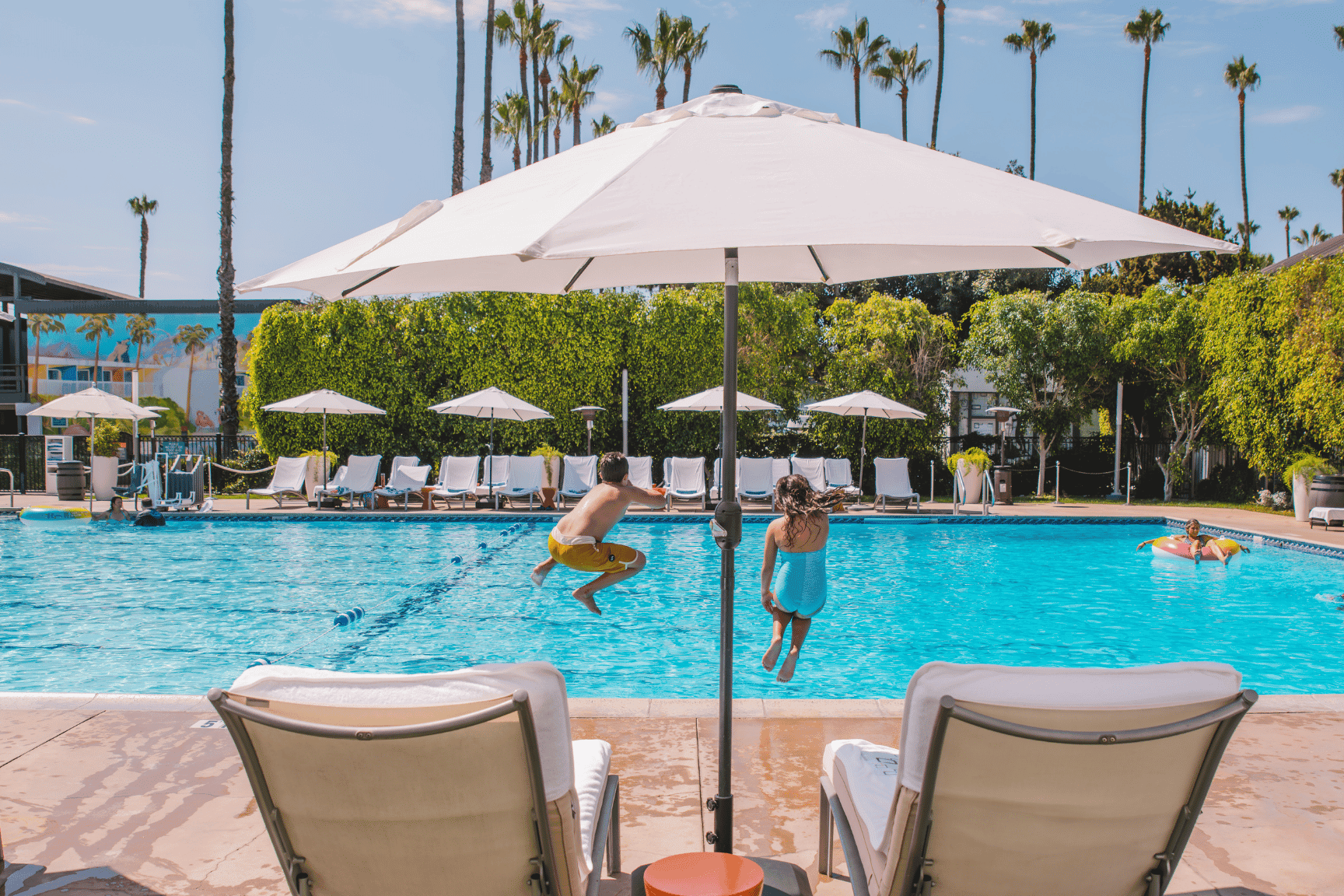 A pool scene with kids jumping in and lounging chairs under umbrellas.