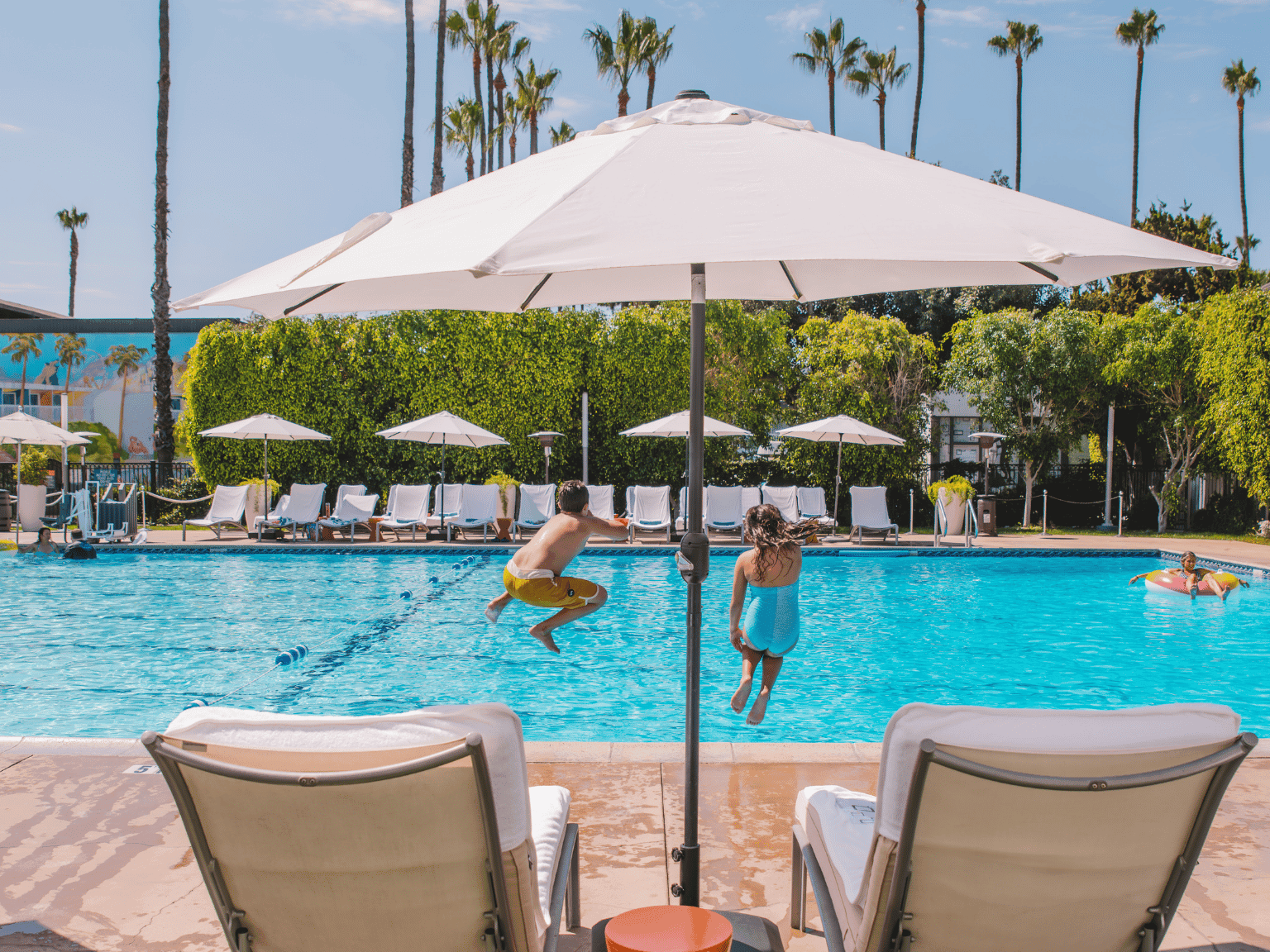 Kids jumping into The Anaheim Hotel Pool with pool chairs and a person in a floatie in the background. 