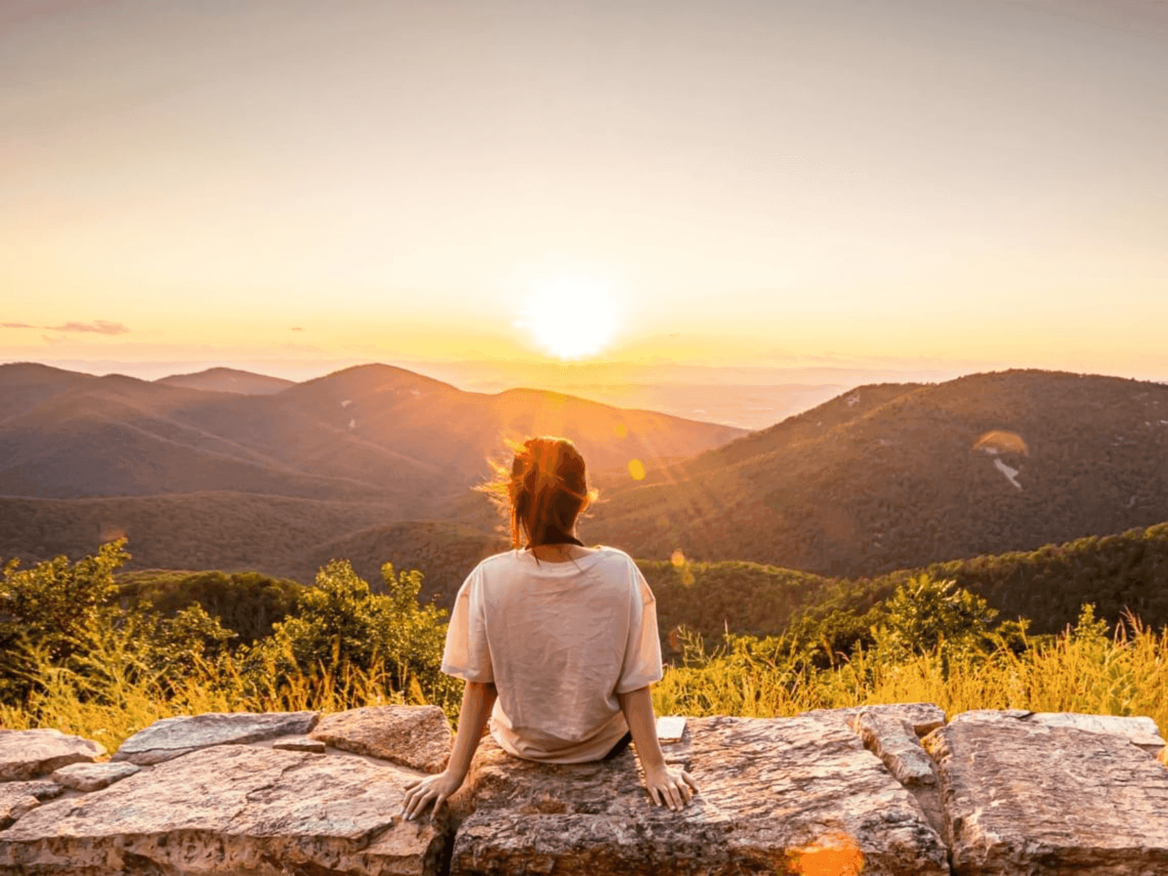 Woman sits on a stone wall, facing sunset over rolling hills and mountains.