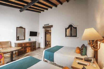 Two beds, wooden table, mirror, dresser, lamp, and chair in a room with white walls and wooden ceiling.