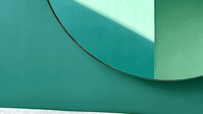 Oval mirror reflecting sunlight above a white velvet couch against a green wall.