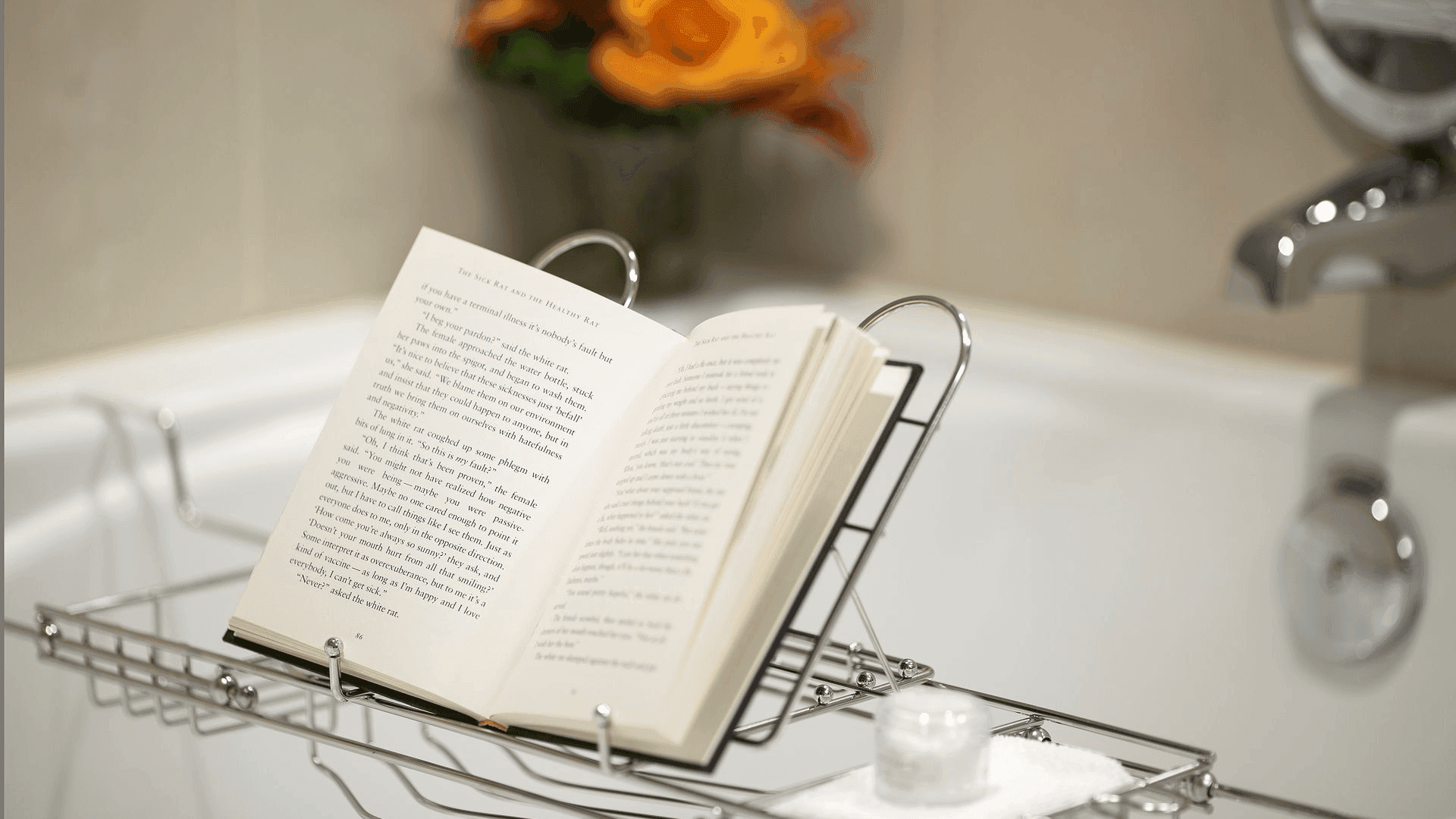 An open book sits on a metal bookstand in a bathtub, with a lit candle and flowers nearby.