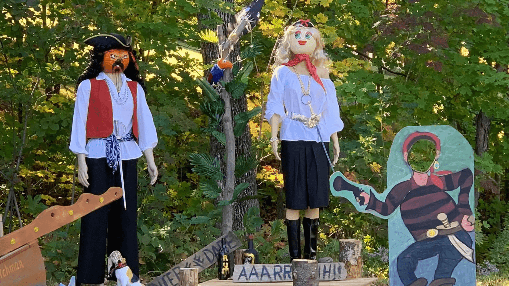 Two pirate mannequins made of pumpkin, a painted pirate board, and props on a wooden platform set in a forest.