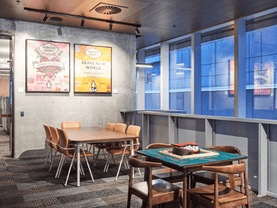 A room with two tables, chairs, two framed posters on wall, and large windows with blue view.