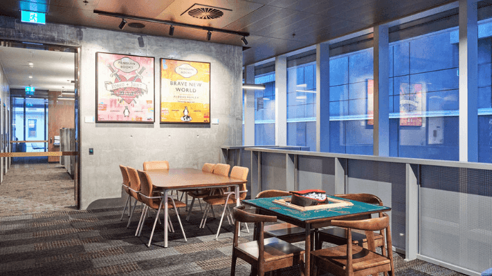 A room with two tables, chairs, two framed posters on wall, and large windows with blue view.