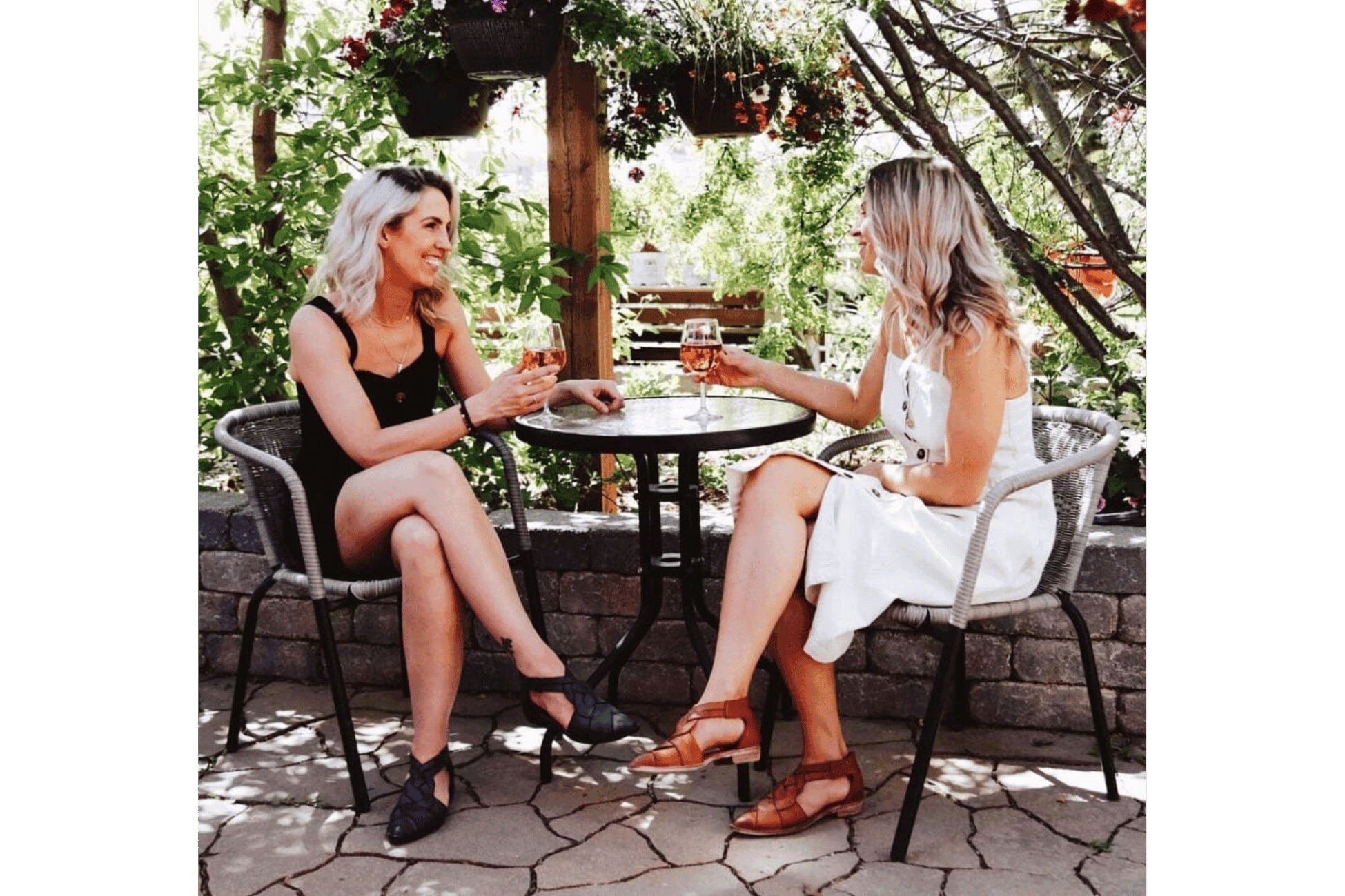 Photograph of two women sitting at a round outdoor table, clinking glasses of wine in a garden setting. Surrounding elements include hanging plants, stone wall, and leafy trees, creating a relaxed and social atmosphere.