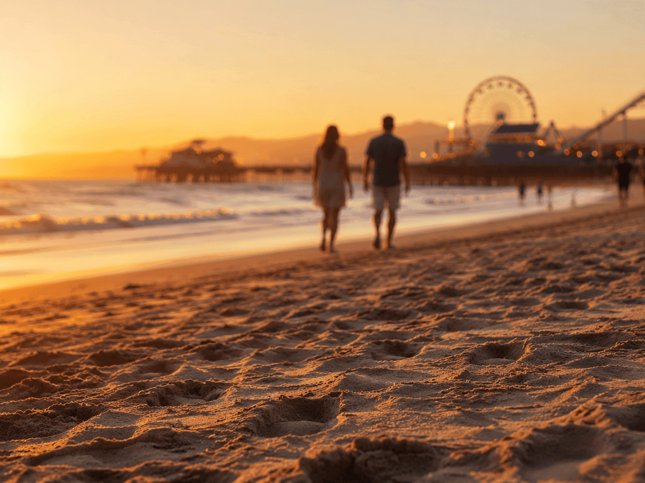 Couple walking along sandy beach at sunset with pier and Ferris wheel visible in background for Summer Begins by the Sea!