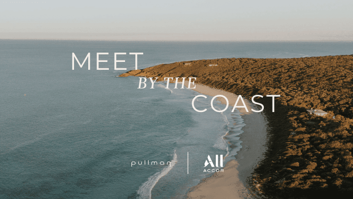 Aerial view of a sandy beach meeting calm ocean waters with rocky coastline and Meet by the Coast text overlay.