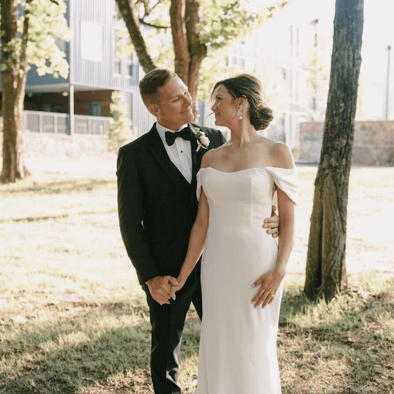A couple in wedding attire stands in a park, holding hands and smiling.