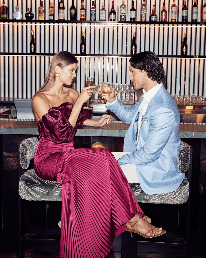 Elegant couple enjoying cocktails at a stylish bar, toasting in a modern setting with upscale décor and vibrant ambiance.