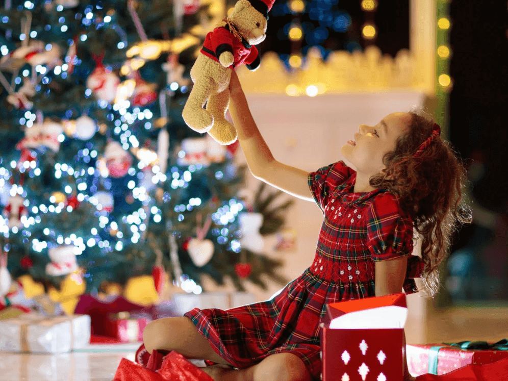 Young girl in a red plaid dress joyfully holding up a teddy bear near a brightly lit Christmas tree at Spring Creek Resorts