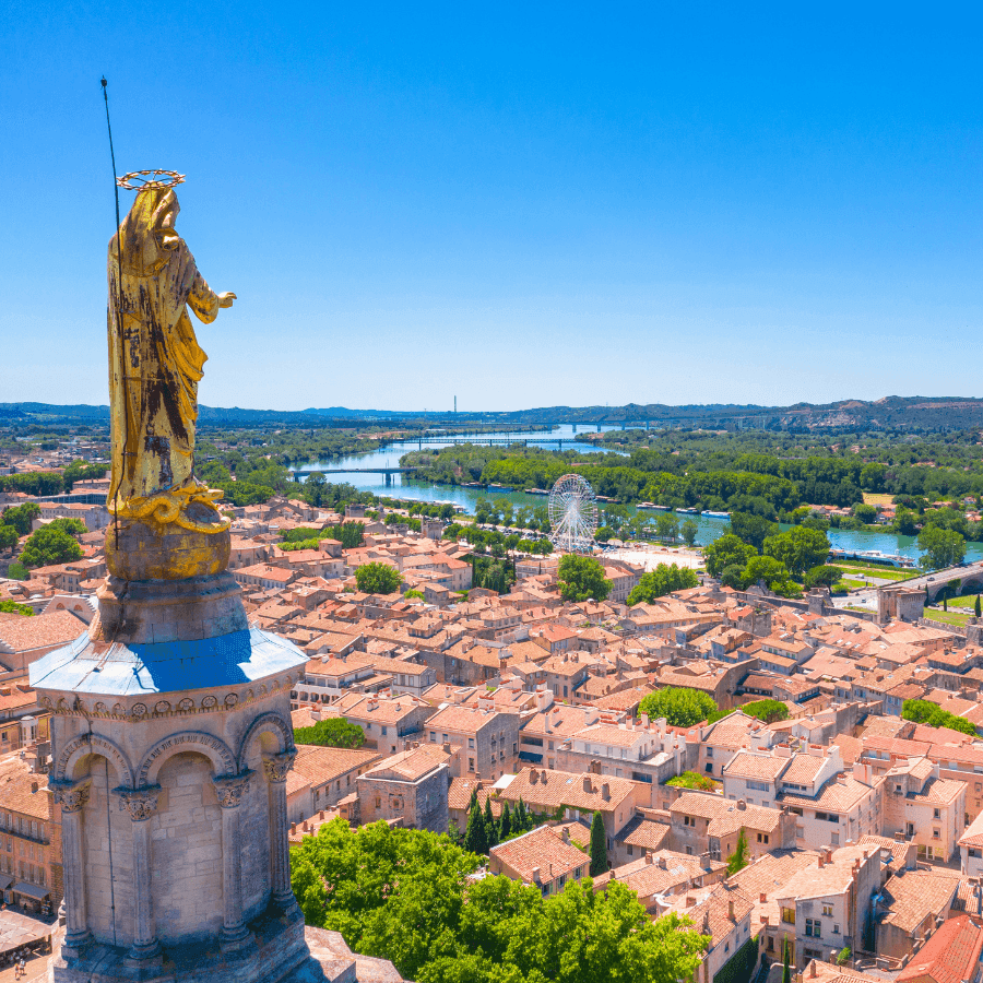 Golden statue atop a church overlooking a sunny cityscape with terracotta roofs & lush greenery near ResidHotel Grand Avignon