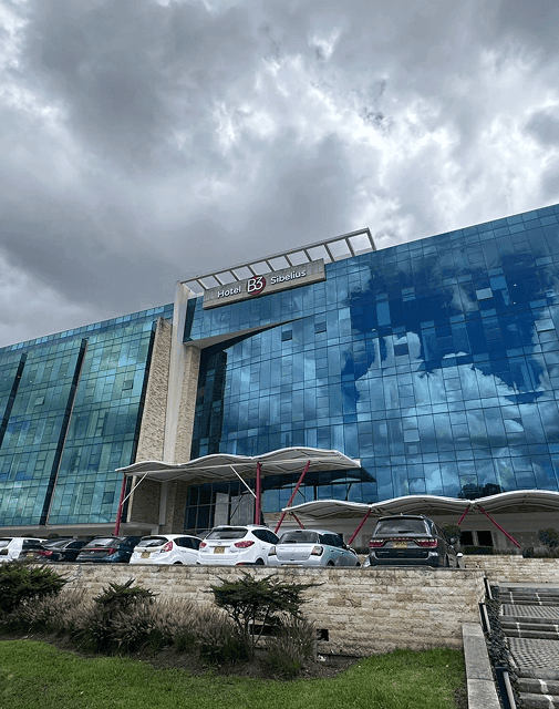Exterior del Hotel B3 Sibelius con paredes de vidrio azul sobre autos estacionados bajo un cielo nublado en Bogotá, Colombia