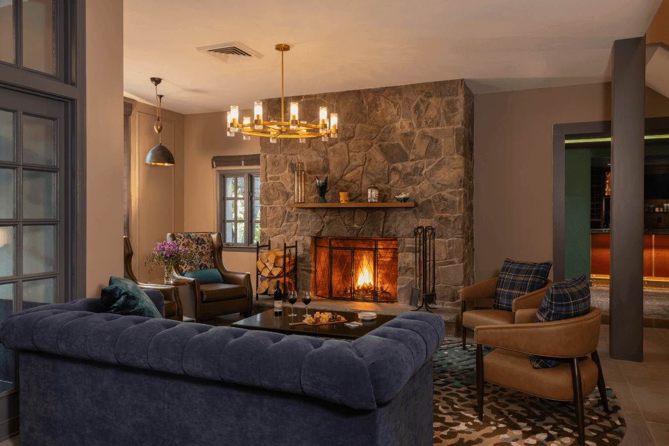 Lounge with a blue velvet sofa and leather chairs by a stone fireplace under a brass chandelier at La Tourelle Inn Spa
