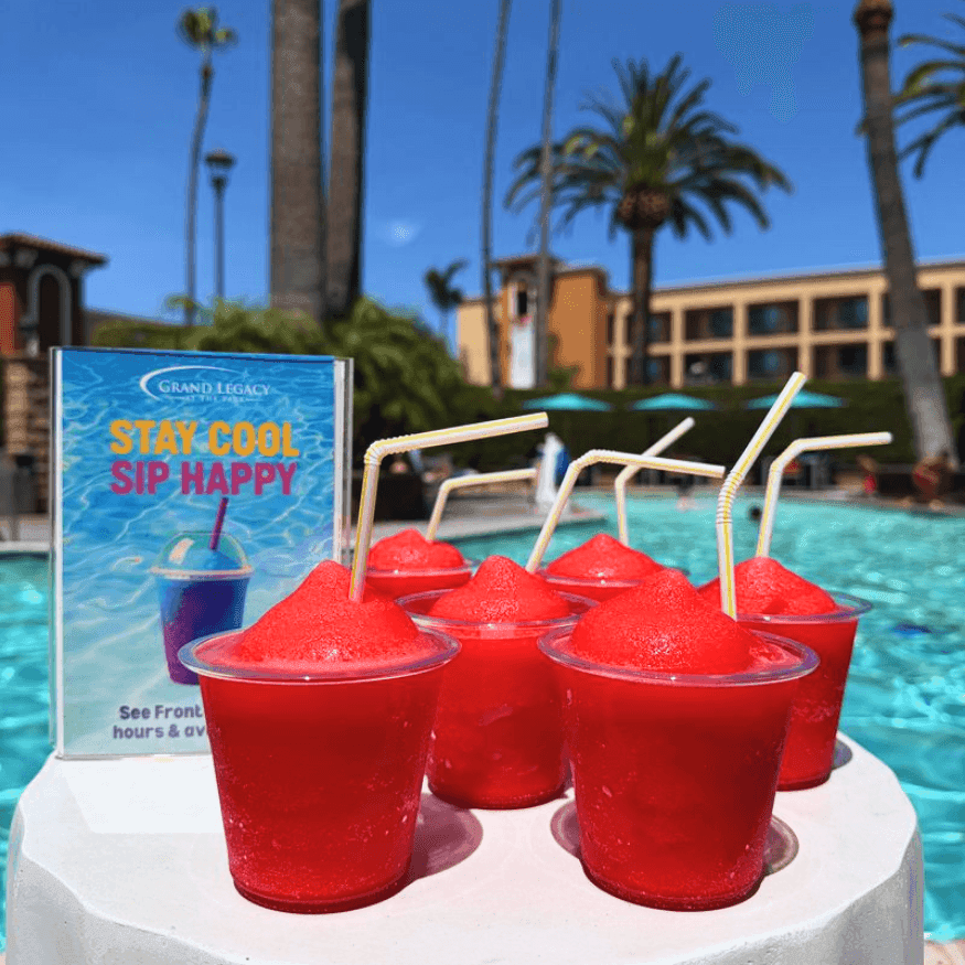 Five red slushies with straws displayed on a white table beside a sparkling pool surrounded by palm trees and resort buildings.