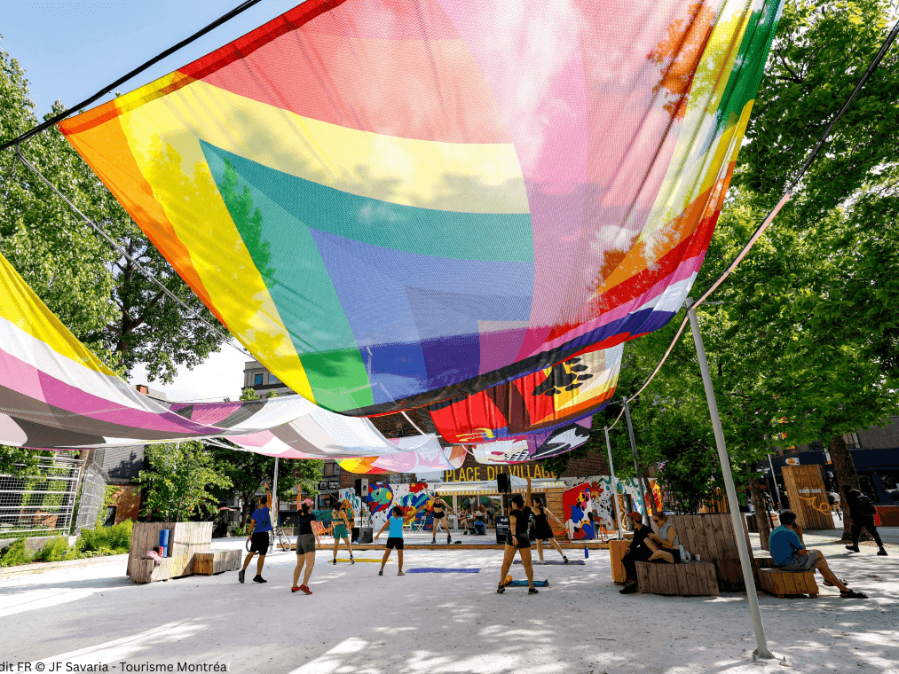Des personnes sous un drapeau arc-en-ciel dans un espace extérieur animé au Village.