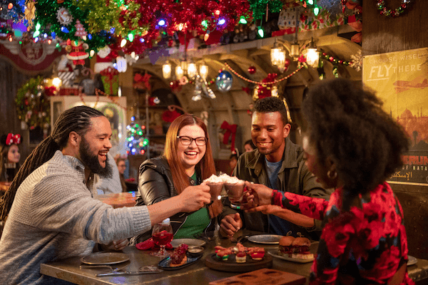 Two couples at a restaurant table toast with hot chocolate beneath colorful Christmas decorations. 