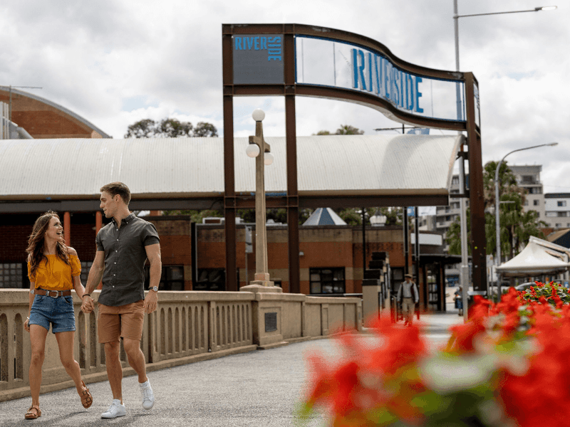 Couple walking hand in hand under a metal sign reading Riverside, with red flowers in the foreground.