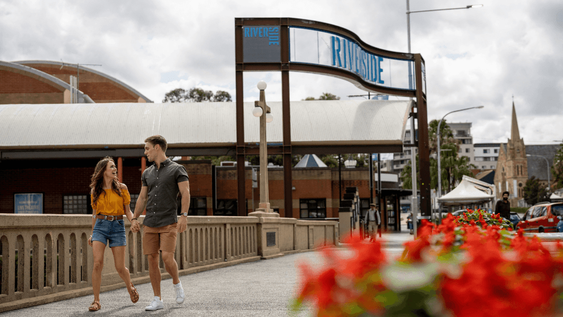 Couple walking hand-in-hand on a bridge with flowers and a riverside sign in the background.