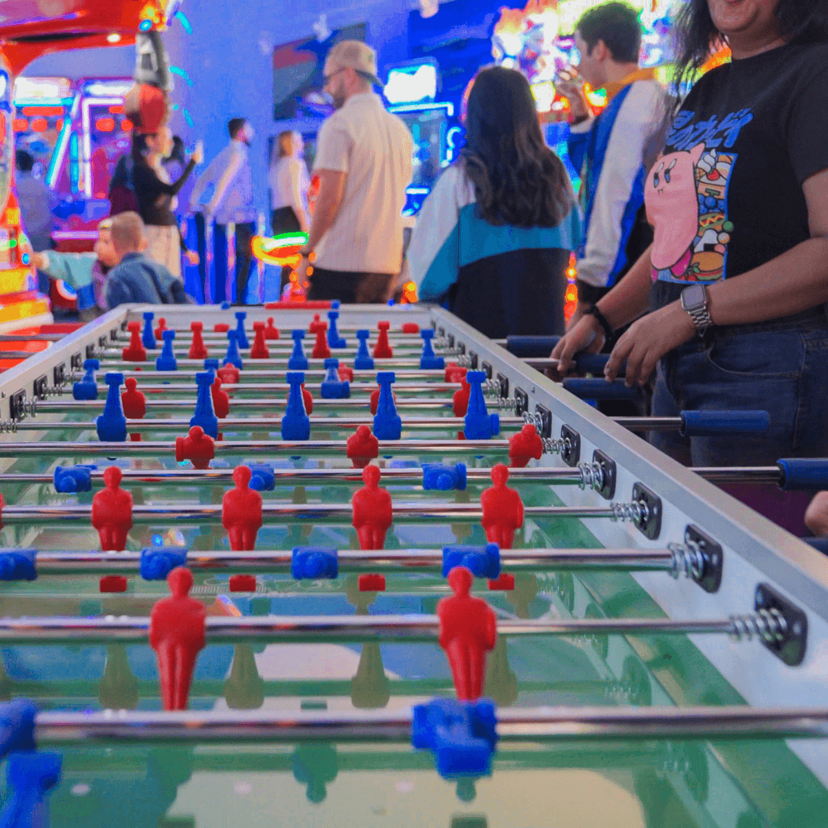 Foosball table with red and blue players, guests gathered nearby under colorful neon arcade lights at Hotel X Toronto