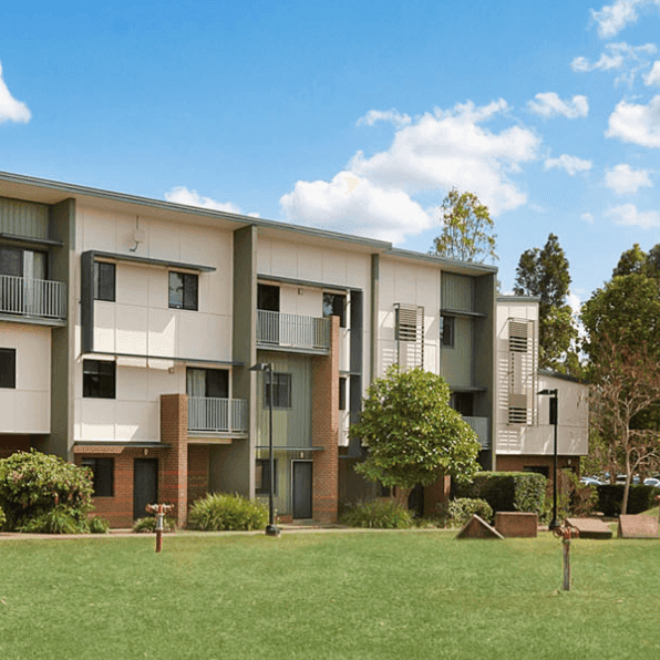 Modern three-story apartment buildings with balconies surrounded by green lawn and mature trees under a blue sky.