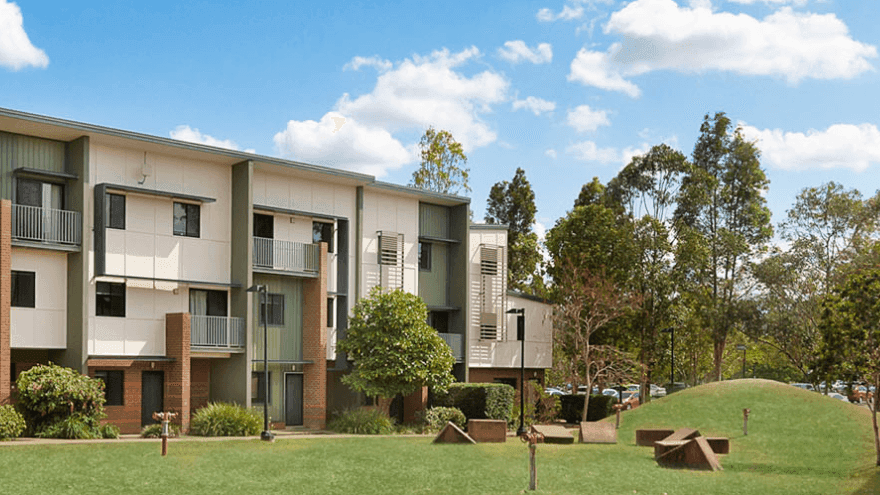 Modern three-story apartment buildings with balconies surrounded by green lawn and mature trees under a blue sky.