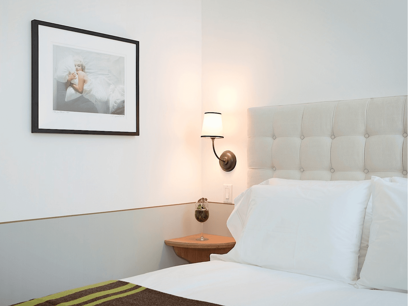 A neatly made bed with white linens and a tufted headboard, with a bedside lamp and framed art on the wall.