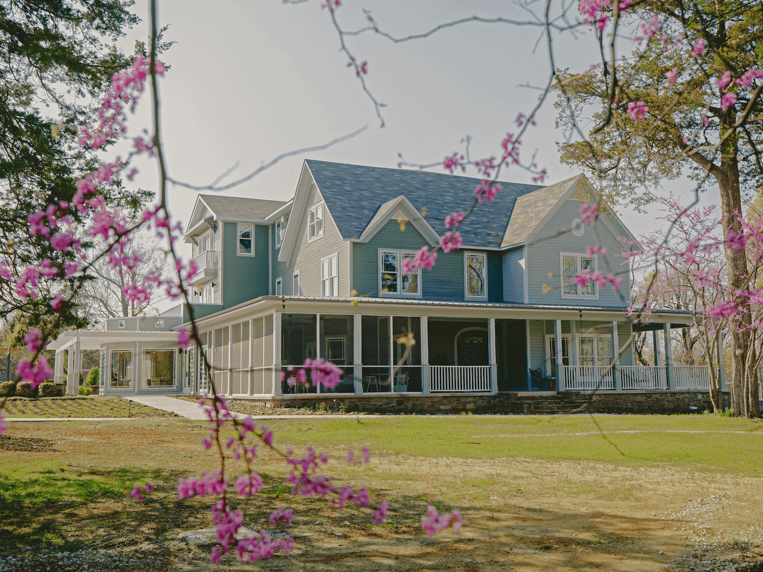 Large blue hotel with a porch, surrounded by trees with pink blossoms and a green lawn.