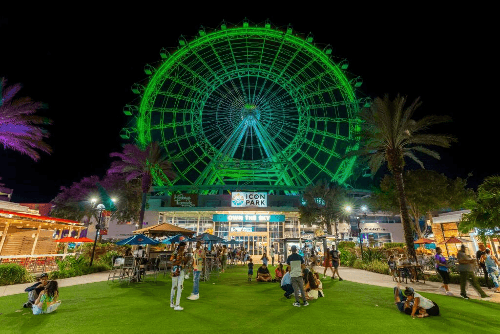 A large, green Ferris wheel glows in the distance at night above a courtyard with green grass surrounded by restaurants and businesses.