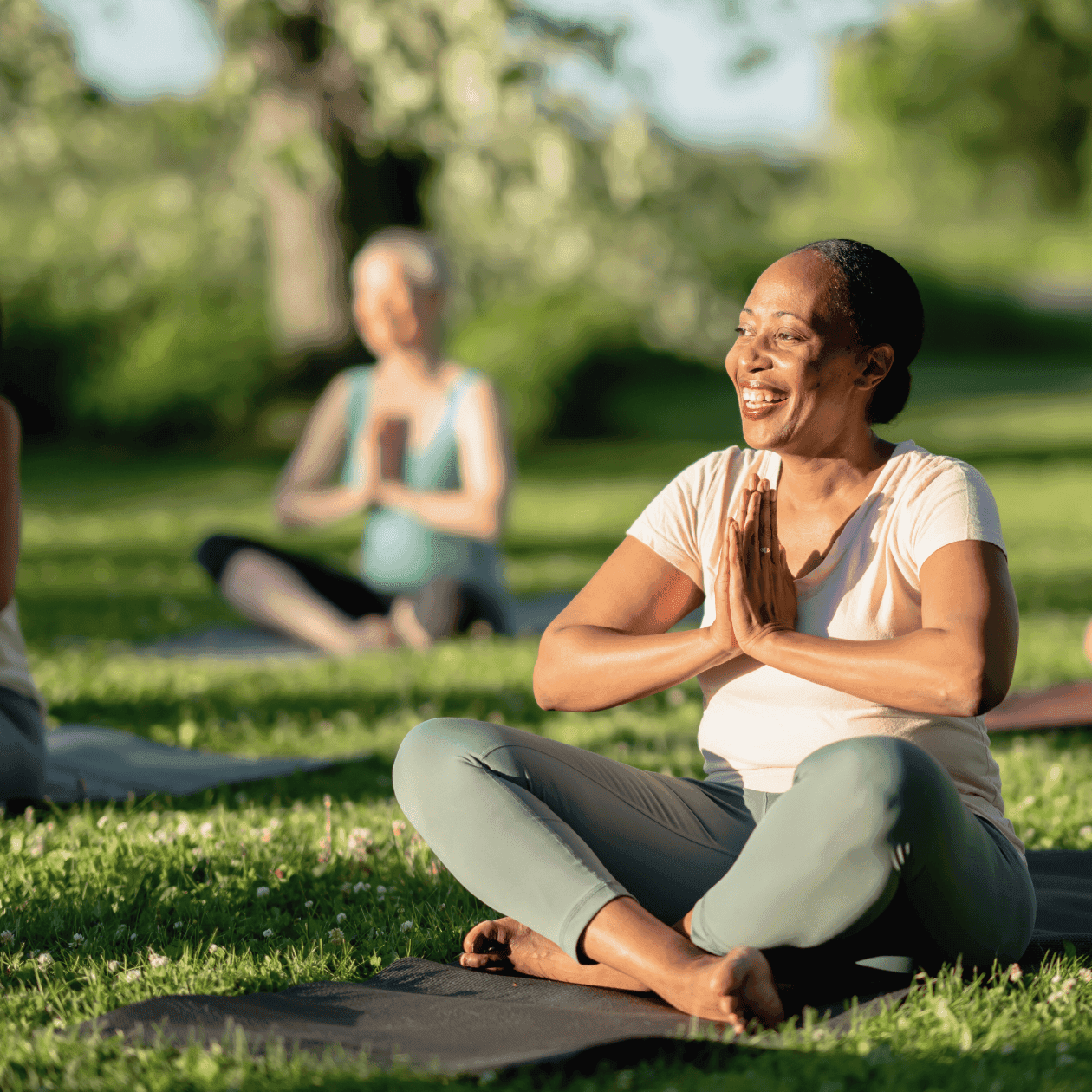 Group of people practicing yoga on a grassy field with one woman smiling and hands in prayer position.