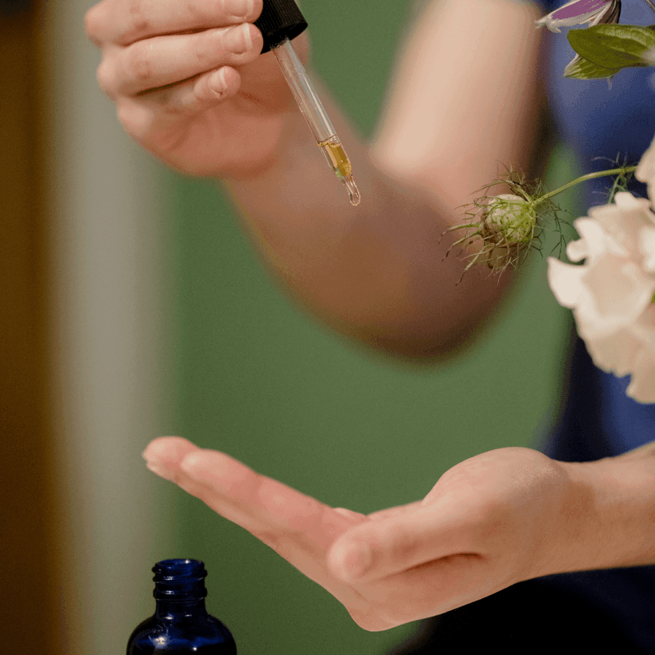 Dropper releasing golden oil into an open palm, beside a blue glass bottle and white blossoms at La Tourelle Inn & Spa