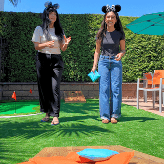 Two women wearing Mickey Mouse ears stand on artificial turf playing cornhole with colorful boards and bean bags outdoors