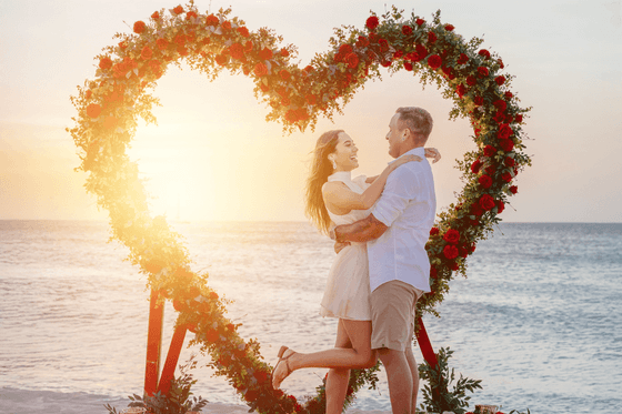 Couple embraces under a heart-shaped floral arch at sunset on the beach.