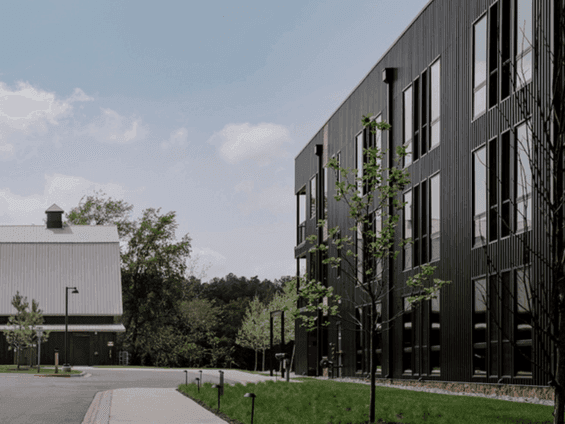 A modern black building with windows next to a grassy area and a gray barn under a blue sky.