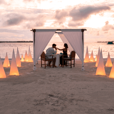 A romantic beach dinner setup with a couple and lit candles at dusk.