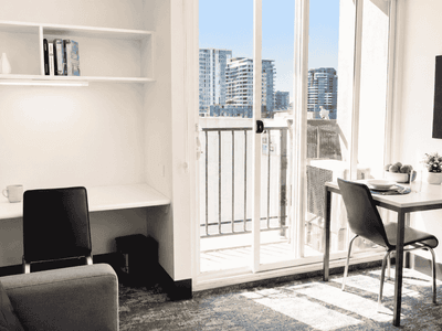 Modern student living space with desk, chair, built-in shelving, and balcony overlooking city skyline at Student Living - Waymouth