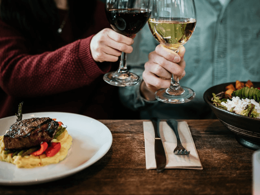 Two people toasting with wine glasses over a table with steak and salad.
