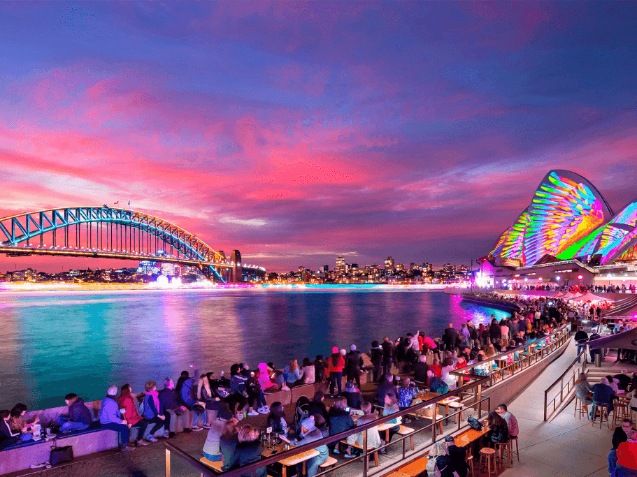 Crowds gather by illuminated Sydney Opera House and Harbour Bridge at dusk during Vivid Sydney 2026.