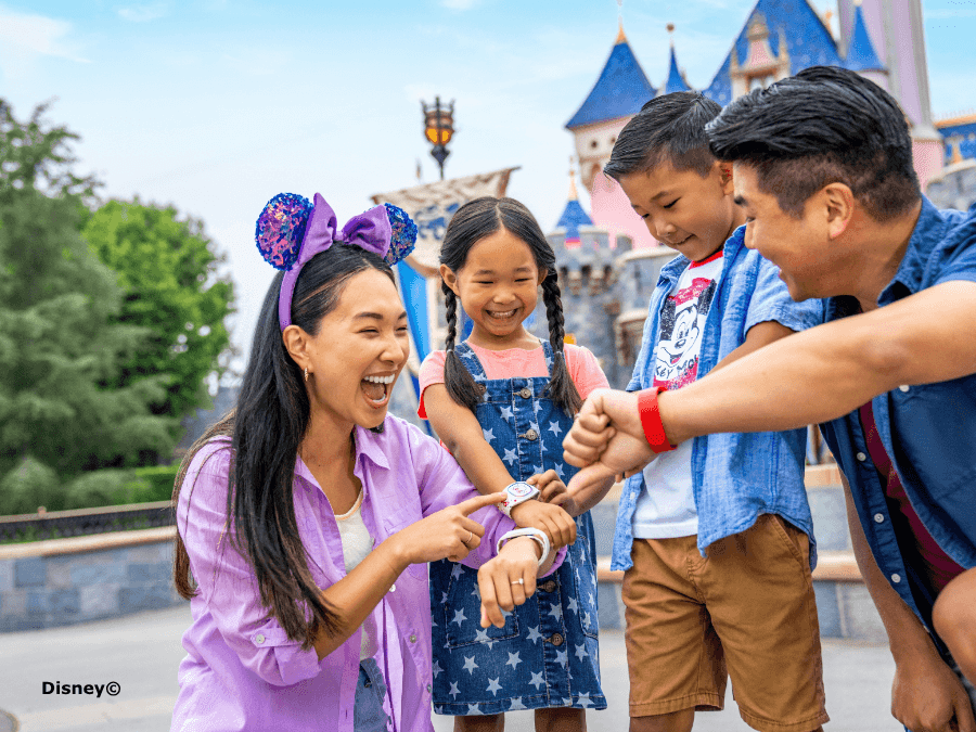 Family of four smiling together while checking wristbands in front of a fairytale castle at a theme park.